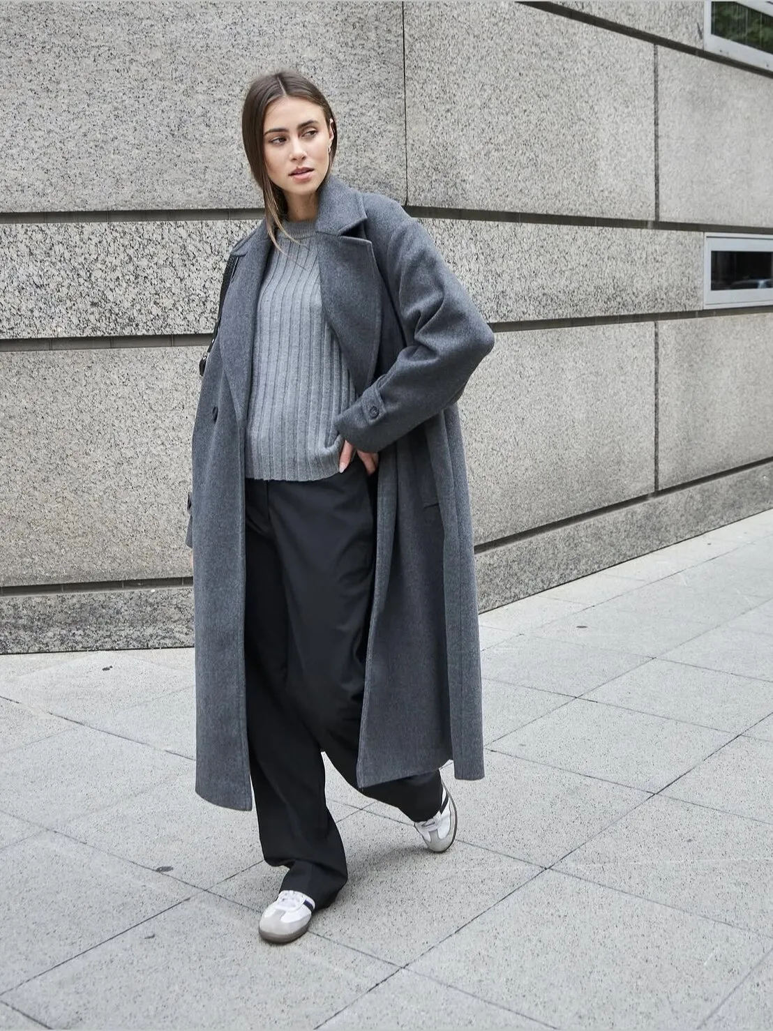 Look urbano con tapado gris largo, sweater acanalado y pantalón negro frente a una pared de piedra.