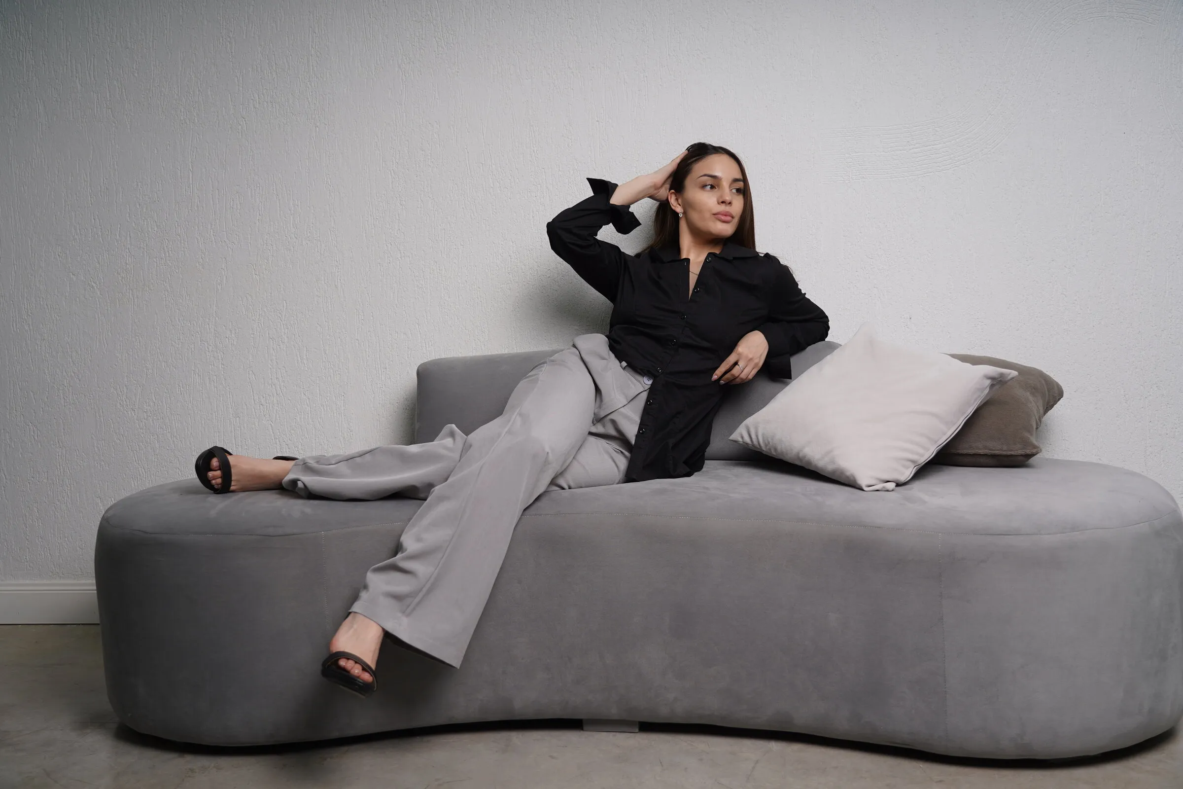 Modelo con camisa negra y pantalón gris recostada en un sillón curvo de pana gris.